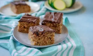 Saftiger Zucchini Kuchen mit Schokoladenglasur auf einem Teller, daneben ein paar Zucchinischeiben.