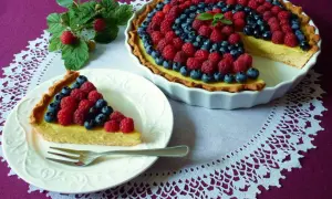 Pyszna tarta z kremem budyniowym, ozdobiona malinami i borówkami. Jeden kawałek już czeka na talerzyku.