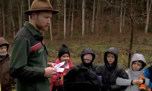 Ein Förster mit braunem Hut erklärt Kindern im Wald etwas. Sie sind von Hütten aus hierher gekommen, um mehr über die Natur zu lernen.