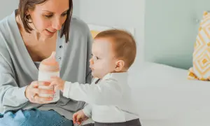 Madre ofrece biberón a su bebé, quien extiende su mano. Momentos tiernos que anticipan las primeras palabras del bebé.