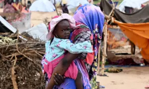 En kvinna bär ett barn i en flyktinglägerliknande miljö. Detta ger en inblick i fakta om Sudan och dess utmaningar.