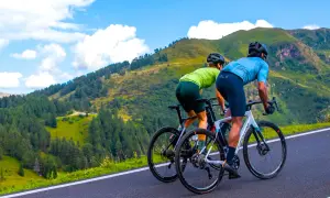 Dwóch kolarzy w sportowych strojach pokonuje górską trasę. Ich pozycja na rowerze szosowym jest aerodynamiczna, gotowi na wyzwanie.