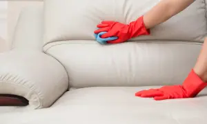 Ręce w czerwonych rękawiczkach delikatnie czyszczą jasne skórzane meble niebieską ściereczką.