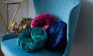 Trzy okrągłe poduszki z weluru w odcieniach turkusu, fuksji i granatu na błękitnym fotelu. Welur wady i zalety: miękkość i elegancja, ale wymaga delikatnej pielęgnacji.