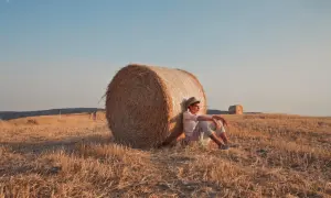 Jonge man met hoed leunt tegen hooibaal in een veld, lijkt verslaafd aan prikkels door de rust.