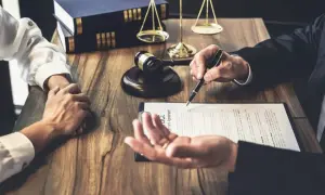 Prawnik wskazuje piórem na umowę, podczas gdy klient trzyma ręce złożone na drewnianym stole. W tle widać młotek sędziowski i wagę sprawiedliwości. To zstępny etap negocjacji.