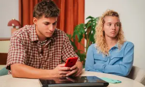 Een jongeman kijkt geconcentreerd naar zijn telefoon, terwijl zijn partner met gekruiste armen en een norse blik wegkijkt. Het voelt alsof hij haar compleet negeert, alsof ze leeggezogen wordt door zijn onoplettendheid.