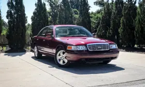 Bordowy Ford Victoria z chromowanym grillem stoi na betonowym podjeździe, otoczony przez wysokie, ciemnozielone drzewa.