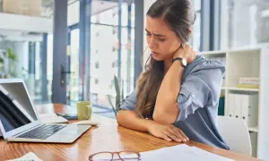 Kobieta z chorobliwym lękiem przed zmianami i wszelkimi nowościami masuje obolałą szyję, siedząc przy biurku z laptopem.