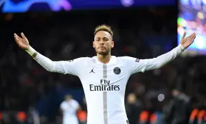 Neymar w białej koszulce PSG, z charakterystycznymi fryzurami, unosi ręce na tle tłumu.
