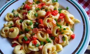 Pyszne tortellini co to jest? Makaron z kawałkami szynki, papryki i szczypiorku na talerzu.