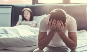Mężczyzna z rękami na twarzy, siedzący na łóżku, czuje się impotent. Kobieta leży obok, zamyślona.