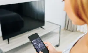 Kobieta trzyma smartfon z aplikacją do sterowania TV, obok stoi włączony telewizor.