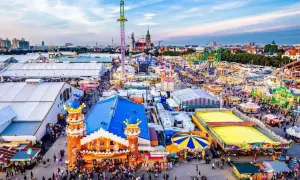 Vista aerea di un'enorme festa Oktoberfest con giostre, bancarelle e folla festante sotto un cielo azzurro.