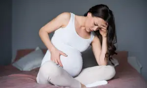 Mujer embarazada sentada en la cama, con la mano en la frente y el vientre, expresando dolor de costillas durante el embarazo.