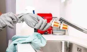 Mani con guanti grigi si preparano a sturare il lavandino, con un panno azzurro e una macchinetta del caffè rossa sullo sfondo.