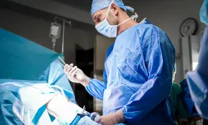 Chirurg w niebieskim fartuchu i masce operacyjnej trzyma narzędzia podczas zabiegu.