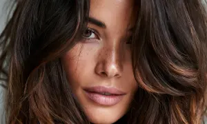 Primo piano di una donna con capelli castani e colpi di sole luminosi, lentiggini sul naso e occhi penetranti.