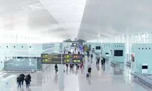 Jasny, nowoczesny terminal lotniska w Hiszpanii. Pasażerowie z bagażami przemieszczają się pod wielkim, białym sufitem, kierując się znakami.