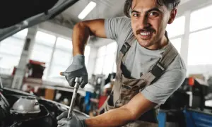Mechanik w ogrodniczkach i rękawiczkach naprawia silnik samochodu kluczem nasadowym.