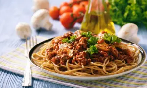 Une assiette de tagliatelles à la bolognaise, garnie de persil frais. Des champignons, tomates et une bouteille d'huile d'olive sont en arrière-plan. Une recette parfaite pour un repas réconfortant.