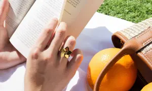 Mano con un anillo dorado con forma de flor, leyendo un libro junto a naranjas y un bolso de mimbre. Las tallas de anillos perfectas para cada ocasión.
