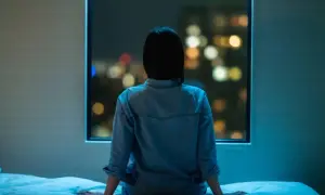 A person sits on a bed, looking out at city lights, feeling the weight of anxiety even when sleeping next to someone.