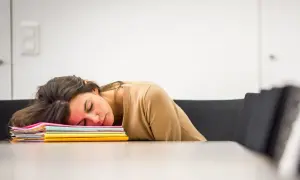 En kvinna sover uttråkad på jobbet, med huvudet vilande på en hög med färgglada mappar.