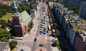 Widok z lotu ptaka na miasto z kościołem i długimi ogrodami. Ulicą jedzie autobus, a obok parkują samochody.