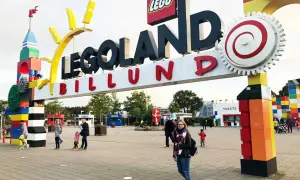 Uśmiechnięta dziewczyna z plecakiem przed wejściem do Legolandu. W tle widać bramę z napisem "LEGOLAND BILLUND" i budki z biletami.