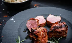 Saftige schweine medaillons, perfekt gegrillt, liegen auf einem dunklen Teller, garniert mit Rosmarin. Daneben eine Schüssel mit cremiger Beilage.