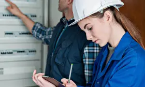 Technicy podczas odbioru instalacji elektrycznej. Kobieta w kasku notuje uwagi, mężczyzna wskazuje na rozdzielnicę.