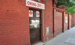 Czerwona ceglana ściana z napisem "OKNO ŻYCIA" i drzwiami. To miejsce daje szansę na nowe życie.