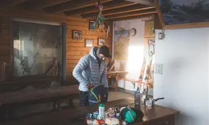 Dans une cabane de falaise, un skieur prépare son sac. Des skis, des cartes et des provisions sont visibles.