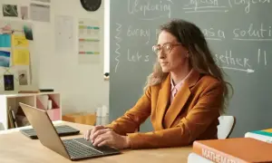Nauczycielka pracuje przy laptopie. Na tablicy widnieje napis "English". Karta oceny pracy nauczyciela może być wypełniana cyfrowo.