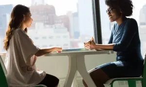 Dos mujeres conversan animadamente en una mesa junto a una ventana. Una de ellas, con gafas, sonríe mientras habla, demostrando su habilidad **speaking b1**.