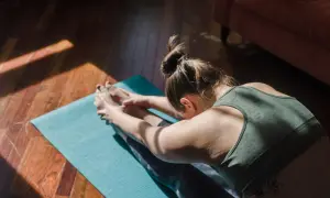 Une femme fait du yoga chez soi, étirant ses jambes sur un tapis bleu, baignée par la lumière du soleil.