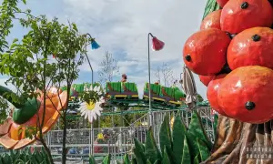 Dzieci cieszą się jazdą w zielonym pociągu-gąsienicy w parku rozrywki. Kolorowe kwiaty i owoce tworzą bajkową scenerię.