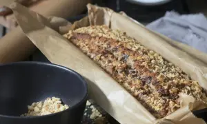 Frisch gebackenes Brot ohne Mehl, bedeckt mit gehackten Nüssen und Schokoladenstückchen, liegt in Backpapier. Daneben eine Schüssel mit Nüssen.