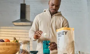 Mężczyzna przygotowuje odżywkę białkową po treningu. Na blacie stoi opakowanie WHEY PROTEIN, shaker i miska z owocami.