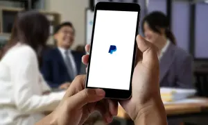 Dłoń trzyma smartfon z logo PayPal na białym ekranie, w tle rozmyte osoby w biurze.
