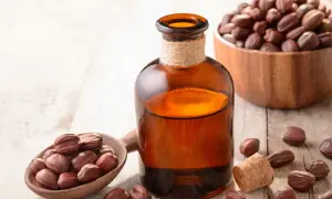 Butelka z bursztynowym płynem, obok drewniana łyżka z nasionami jojoby i miska pełna nasion. Naturalny olej jojoba na włosy.