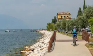 Ciclista sul lungolago, con barche e montagne sullo sfondo. Un assaggio di cosa vedere sul Monte Baldo, tra natura e relax.