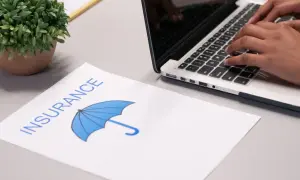 Osoba wpisuje dane na laptopie, obok leży dokument z napisem "INSURANCE" i symbolem parasola, sugerujący ubezpieczenie domu od wszystkich ryzyk.