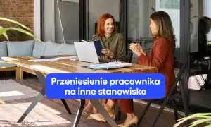 Dwie kobiety rozmawiają przy biurku, omawiając zmianę stanowiska pracy. Jedna z nich trzyma notatnik.