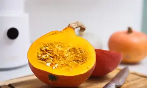 Pomarańczowa dynia przekrojona na pół, pełna nasion, obok inne odmiany dyni i nóż na desce.
