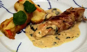 En kycklingrulle insvept i bacon, serverad med en krämig sås till kyckling och gratinerade potatisbitar med gröna blad.
