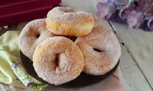 Graffe senza patate, soffici e dorate, ricoperte di zucchero. Un dolce invitante su un piatto scuro, con fiori sullo sfondo.