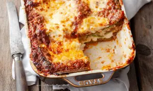 Lasagne alla bolognese appena sfornate, dorate e filanti, in una teglia Staub. Un coltello vintage è appoggiato accanto.