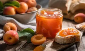 Ein Glas leuchtend orangefarbener Aprikosenmarmelade mit Alkohol, daneben frische Aprikosen und ein Brot mit Marmelade.